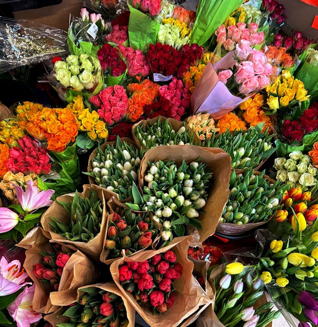 A vibrant display of fresh flowers, including numerous bouquets of tulips, roses, and carnations in various colors such as pink, red, orange, yellow, and white, arranged on a display table. The tulips
