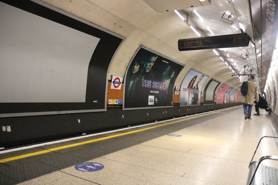 Underground station platform with a curved ceiling and bright strip lighting, featuring multiple advertising posters along the wall, including one for 'The Sister' TV show. A person wearing a beige co