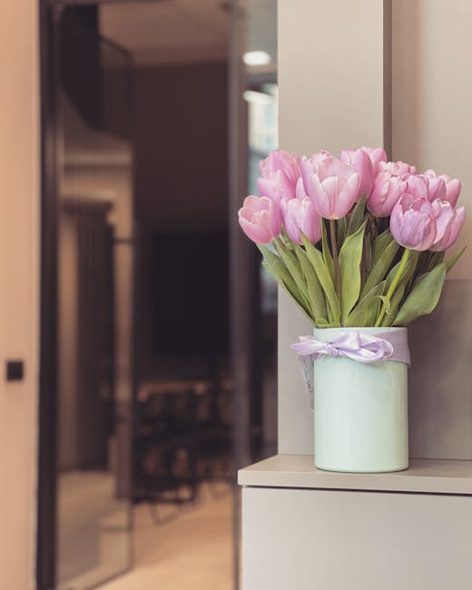 A bouquet of pink tulips with smooth, rounded petals and lush green leaves arranged in a cylindrical, pale green ceramic vase tied with a light purple ribbon. The flowers are fresh, with vibrant petal