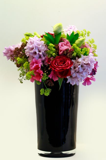 A vibrant floral arrangement in a tall, sleek black vase, featuring a mix of fresh flowers including a central red rose, light pink and pastel purple hydrangeas, white tulips with green buds, and lush
