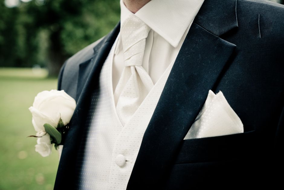 Close-up view of a groom's suit during a wedding, featuring a white rose boutonniere with green leaves pinned to the left lapel of a dark navy tailored jacket. The groom wears a white dress shirt with